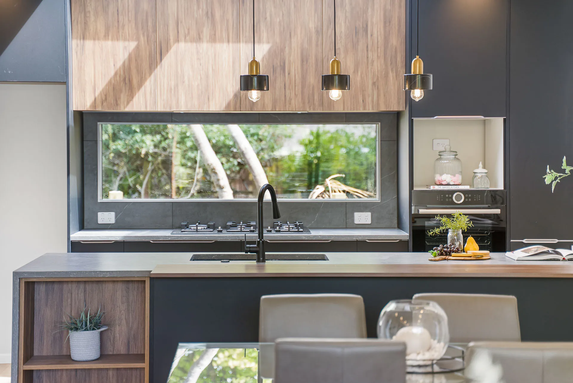 Bucklands Beach kitchen detail, with timber upper cabinets, charcoal cabinetry, three pendant lights and a window above the cooktop.