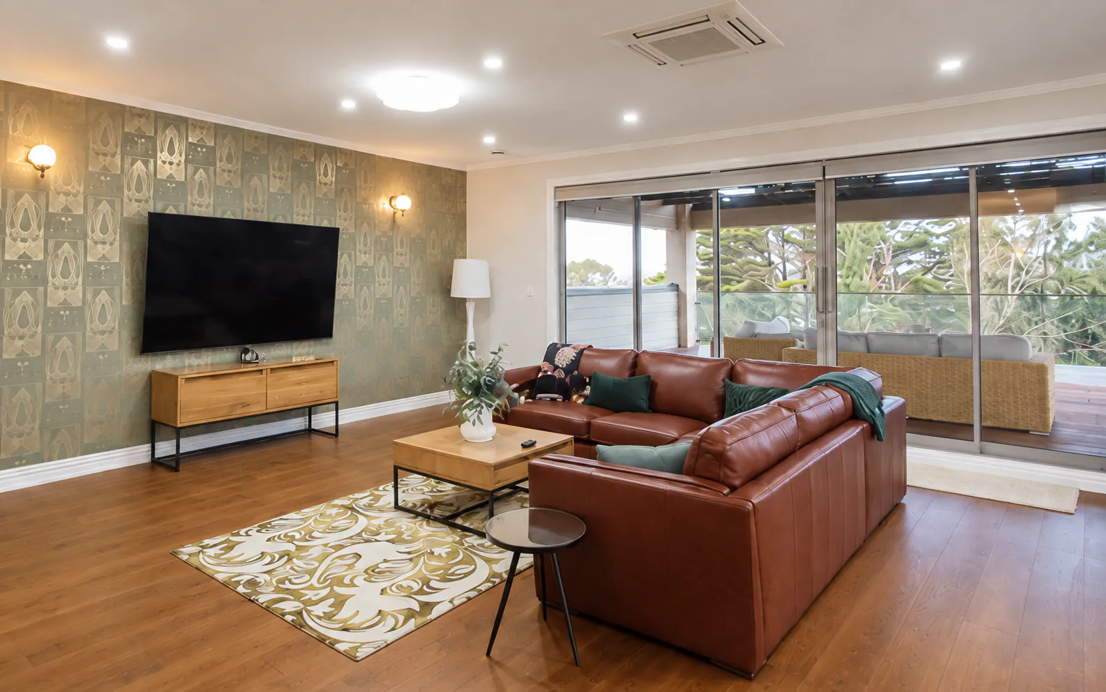 AI-retouched project photograph: the Whitford living room with brown leather sofas, patterned wallpaper, timber floors and doors opening to the terrace.
