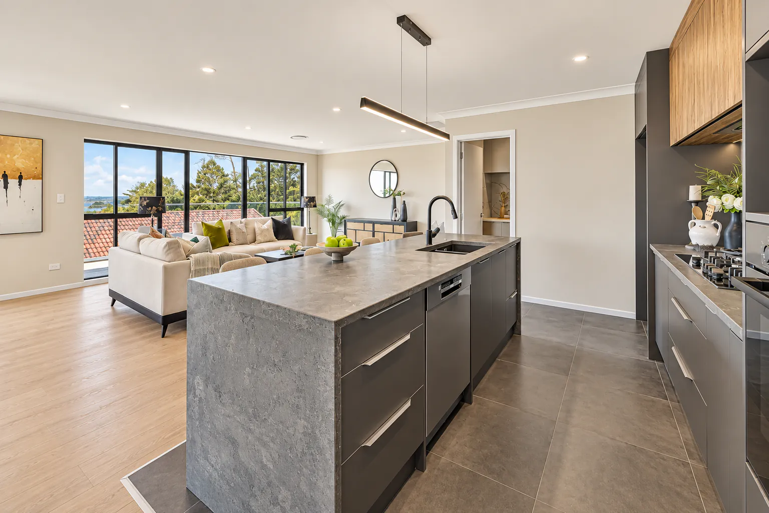 AI-retouched project photograph: the kitchen with a grey waterfall island, charcoal cabinetry and timber upper cabinets at Bucklands Beach 26.