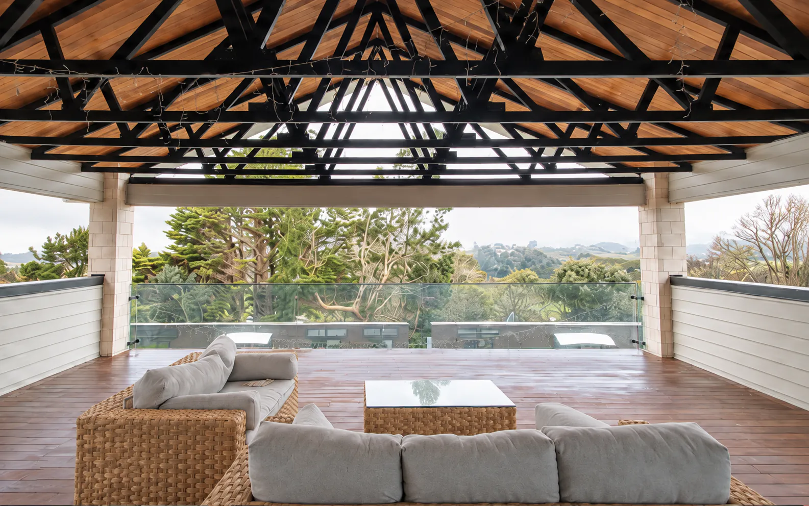 AI-retouched project photograph: the Whitford covered terrace with a timber ceiling, dark exposed trusses, wicker seating and views through a glass balustrade.